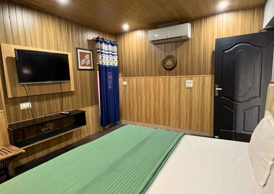 Cozy bedroom with wood-panel walls, wall-mounted TV, AC, and king bed with green duvet at The Basera penthouse.