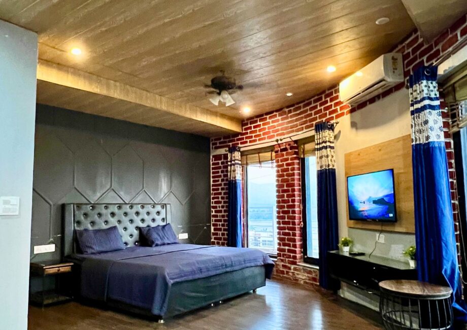 Large suite with dark accent wall, blue king bed, windows with valley view, and TV at The Basera penthouse.