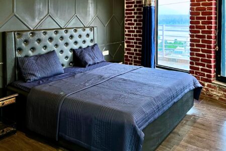Blue upholstered king bed beside floor-to-ceiling windows with Ganga view at The Basera penthouse.