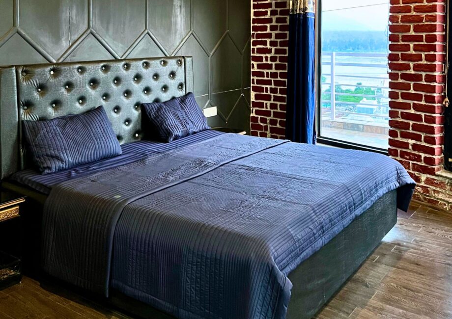 Blue upholstered king bed beside floor-to-ceiling windows with Ganga view at The Basera penthouse.