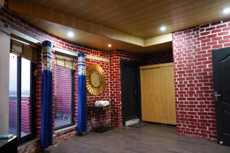 Spacious room corner featuring red brick walls, large blue curtains, a gold sunburst mirror, wooden wardrobe, and warm recessed ceiling lights.