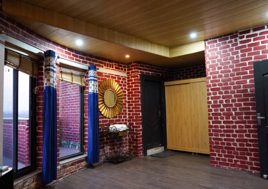 Spacious room corner featuring red brick walls, large blue curtains, a gold sunburst mirror, wooden wardrobe, and warm recessed ceiling lights.