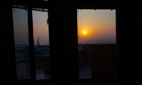 Sunset seen through glass terrace doors, with tiled patio and brick parapet silhouetted against the glowing horizon.