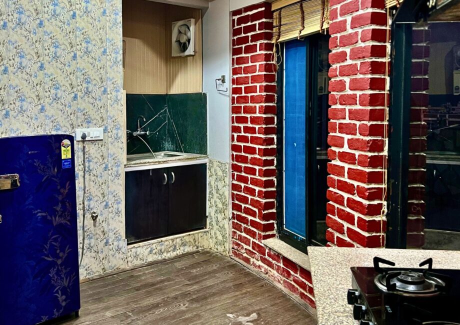 Compact kitchenette with blue refrigerator, floral wallpaper, sink counter and red brick wall by a sliding window.