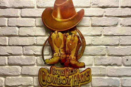 Vintage wall decor of a cowboy hat and boots reading “Boots Chaps Cowboy Hats” on a white brick wall.
