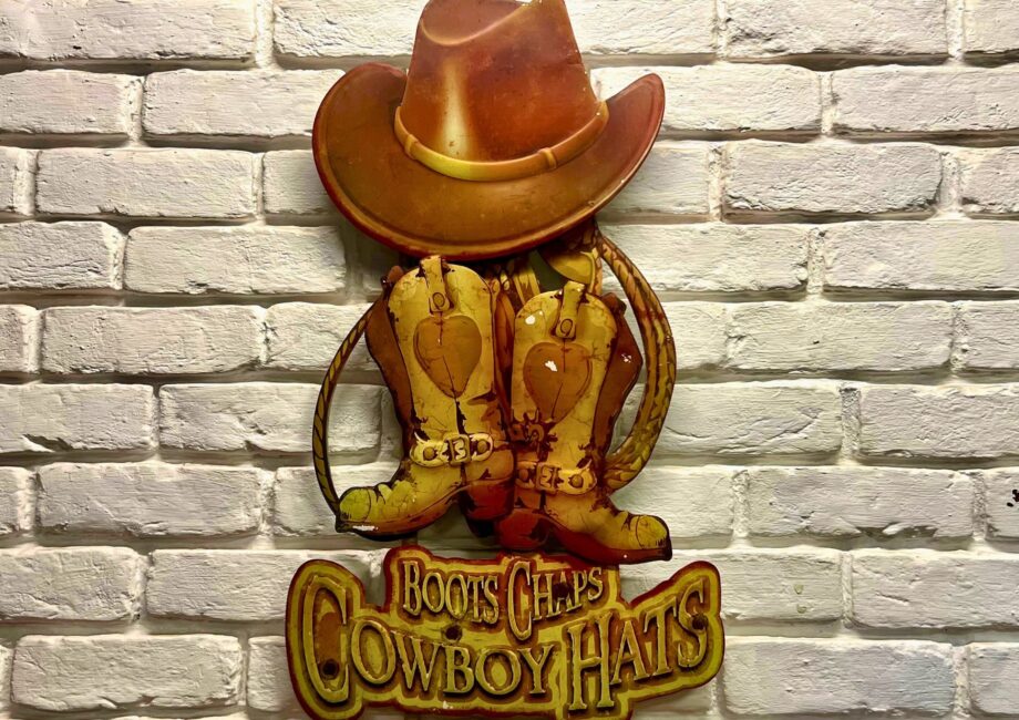 Vintage wall decor of a cowboy hat and boots reading “Boots Chaps Cowboy Hats” on a white brick wall.