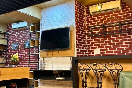 Lounge wall with mounted TV, exposed red-and-white brick, open shelving and three wrought-iron bar stools.