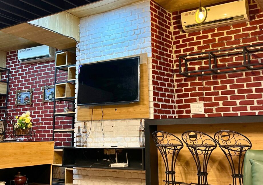 Lounge wall with mounted TV, exposed red-and-white brick, open shelving and three wrought-iron bar stools.
