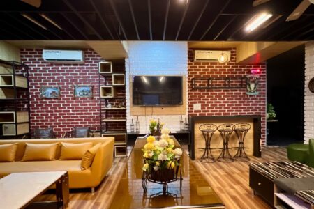 Spacious living area with tan sectional, long glossy table with flower carts, brick walls and a mounted TV.