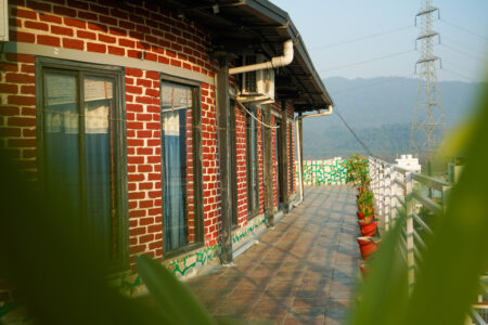 Penthouse terrace walkway with red-brick facade, potted plants, and distant mountain views in Rishikesh