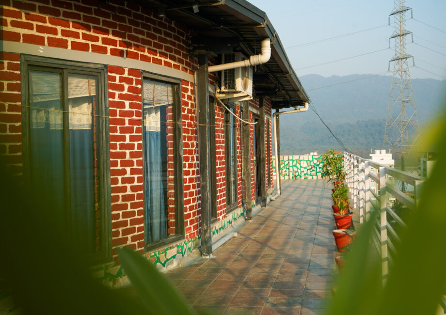 Penthouse terrace walkway with red-brick facade, potted plants, and distant mountain views in Rishikesh