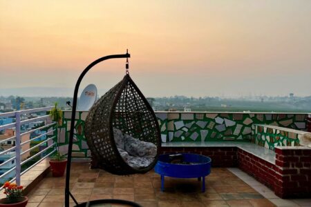 Rooftop terrace at sunset with hanging rattan egg chair, blue fire pit, and wide city–river view.