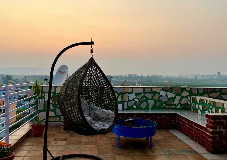 Rooftop terrace at sunset with hanging rattan egg chair, blue fire pit, and wide city–river view.