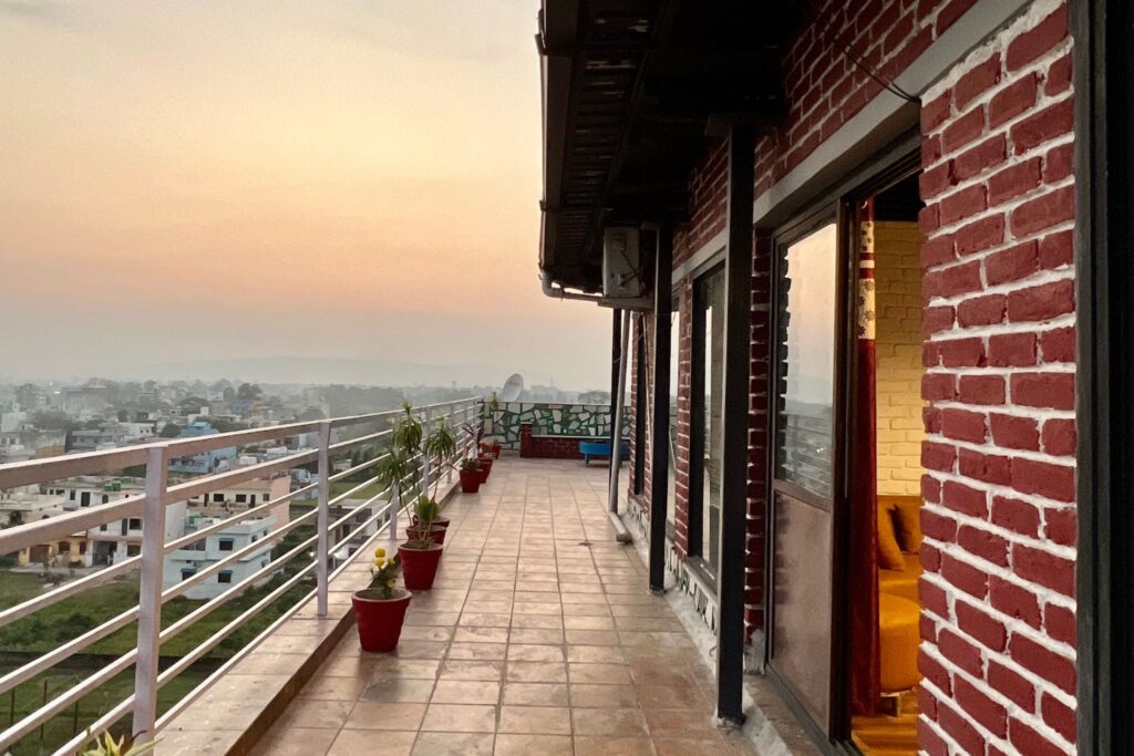 Long wraparound terrace lined with potted plants outside red-brick penthouse, warm evening sky beyond.