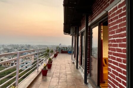 Long wraparound terrace lined with potted plants outside red-brick penthouse, warm evening sky beyond.