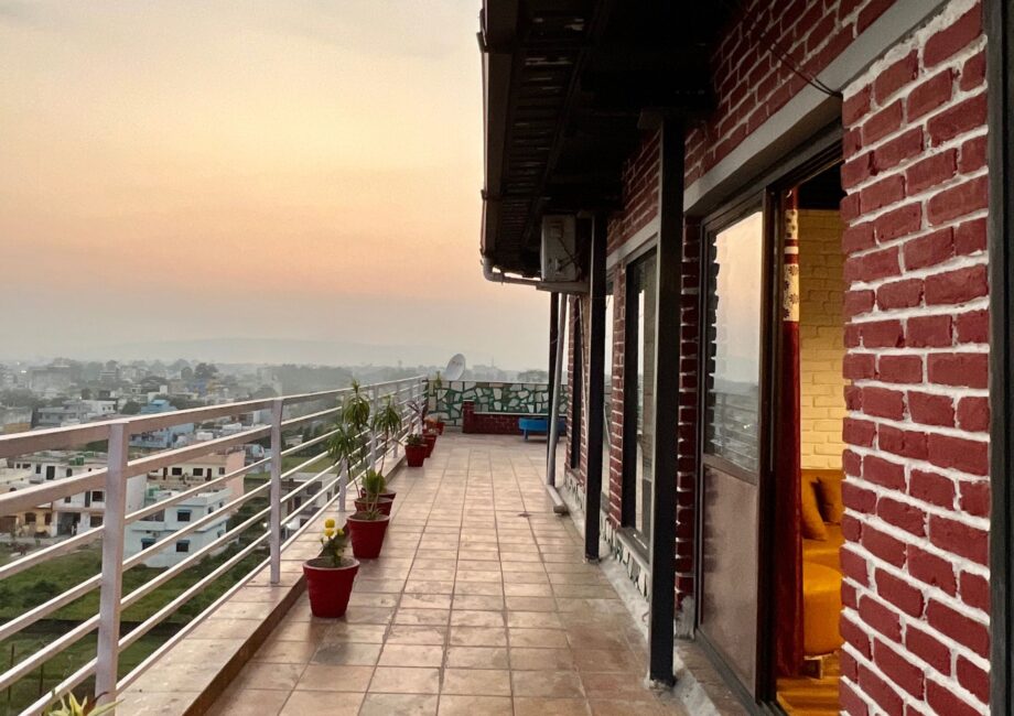 Long wraparound terrace lined with potted plants outside red-brick penthouse, warm evening sky beyond.