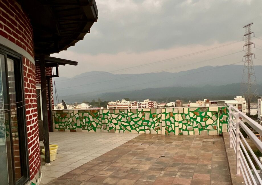 Terrace corner with mosaic parapet, mountain and forest views, and a power pylon under cloudy sky.