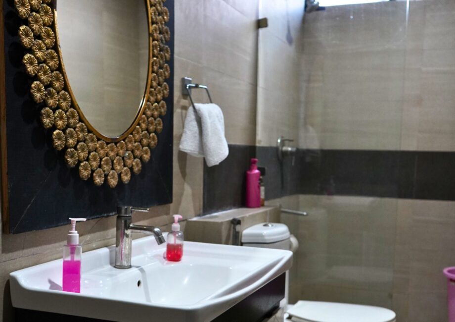 Modern bathroom with round gold-framed mirror, vanity sink, glass shower partition, and toilet at The Basera penthouse.