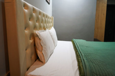 Close-up of tufted beige headboard with white pillows and a neatly folded green quilt against a soft gray bedroom wall.