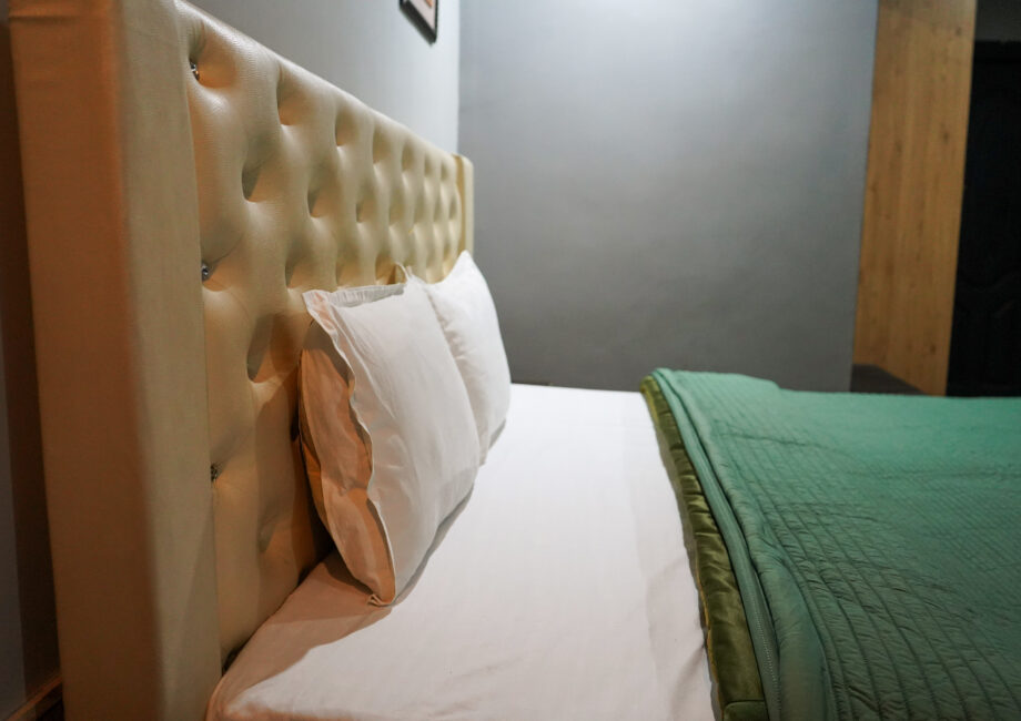 Close-up of tufted beige headboard with white pillows and a neatly folded green quilt against a soft gray bedroom wall.