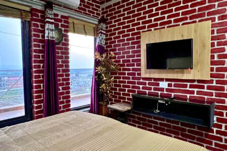 Bedroom with city and Ganga views, red brick walls, wall TV, and purple drapes at The Basera penthouse.