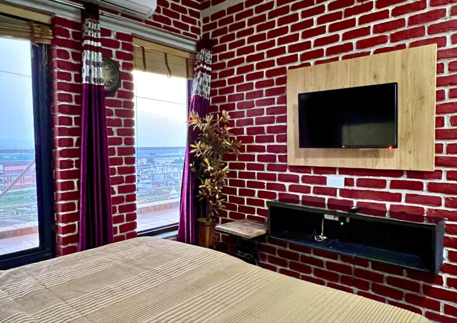 Bedroom with city and Ganga views, red brick walls, wall TV, and purple drapes at The Basera penthouse.