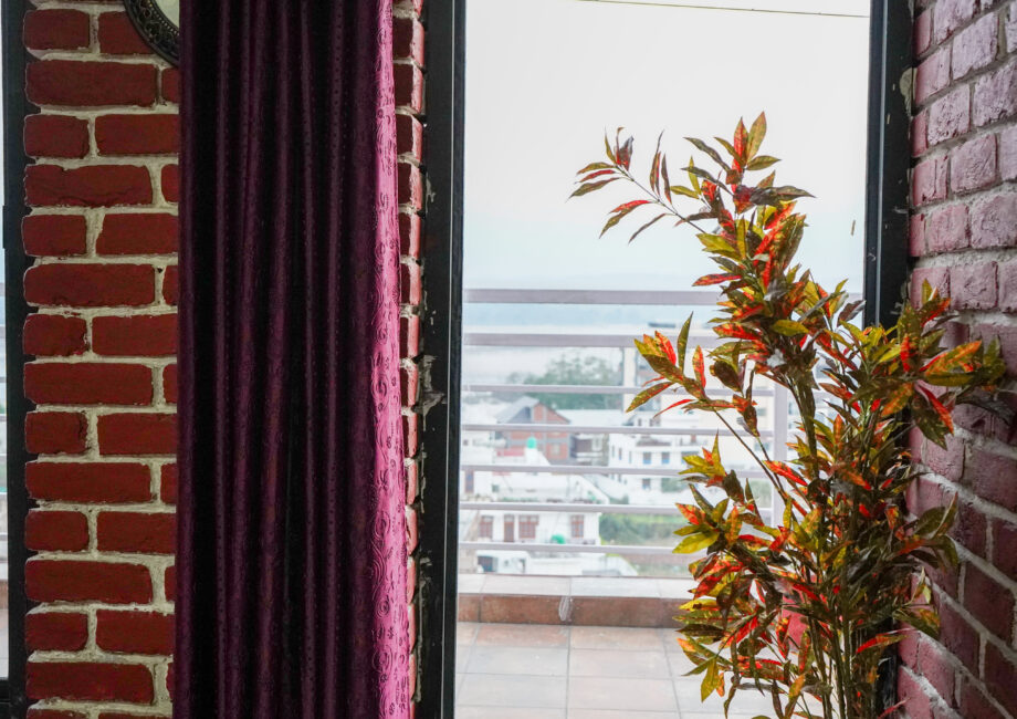 Window nook with purple curtains and a potted plant, opening to a balcony rail and distant city view beyond.