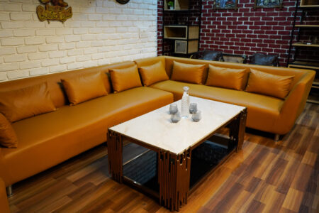 Warm living room with tan L-shaped leather sofa, marble-top coffee table with carafe and glasses, wood floors, whitewashed brick and red-brick accents.