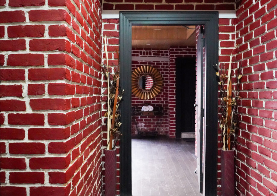 Red-brick hallway framed by a dark door casing, looking into a room with a sunburst mirror and towel console on dark wood flooring.