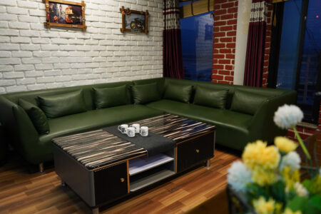 Green sectional sofa with striped black coffee table and mugs in an industrial loft space with white brick wall, red-brick corners, and deep burgundy drapes.