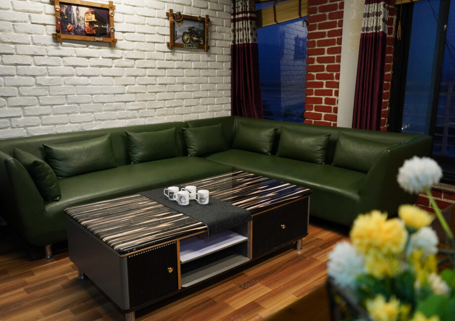 Green sectional sofa with striped black coffee table and mugs in an industrial loft space with white brick wall, red-brick corners, and deep burgundy drapes.